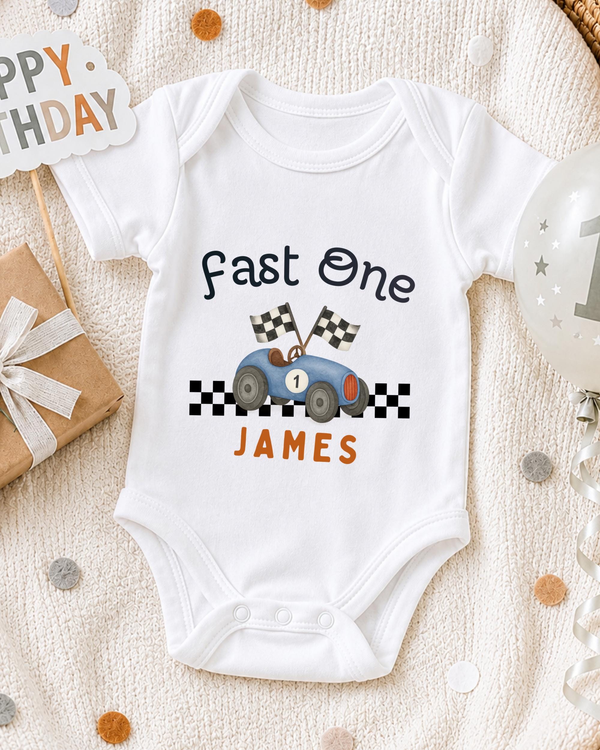 A white onesie with a racing car design and the text "Fast One James" printed on it, along with a birthday sign and a balloon.