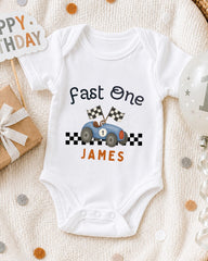 A white onesie with a racing car design and the text "Fast One James" printed on it, along with a birthday sign and a balloon.