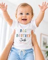A smiling baby wearing a white shirt with the text "BIG BROTHER Jack EST 2023" is being held up by someone's arms.