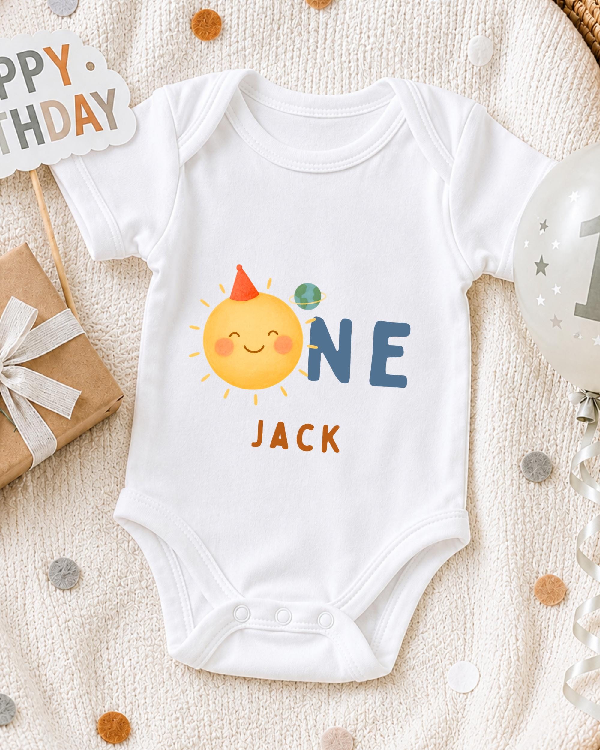 A white onesie with a sun and a party hat on it, along with a "Happy Birthday Jack" sign, is displayed on a bed with confetti.