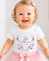 A smiling baby girl wearing a white t-shirt with a floral design and a pink bow in her hair.