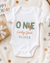 A white onesie with the text "ONE Lucky Duck Oliver" printed on it, along with a birthday sign and a balloon, all placed on a white blanket with confetti.