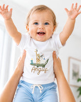 A smiling baby wearing a white t-shirt with a giraffe and the name "Angus" printed on it, being held up by someone's arms.