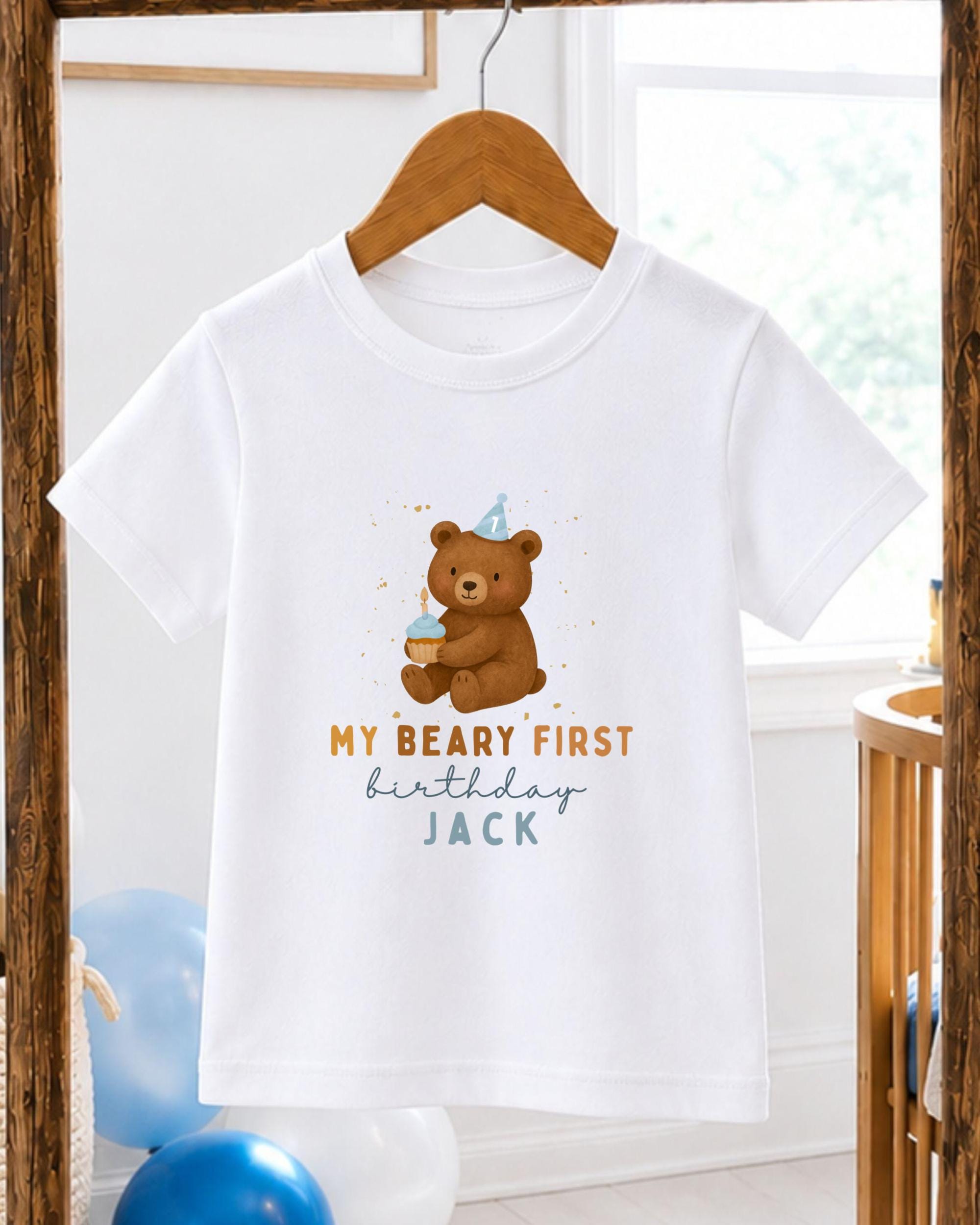 A white t-shirt with a printed design of a teddy bear wearing a party hat and holding a cupcake, along with the text "My Beary First Birthday Jack".