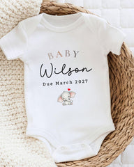 A white baby onesie with the text "BABY WILSON Due March 2027" printed on it, along with an illustration of a baby elephant.