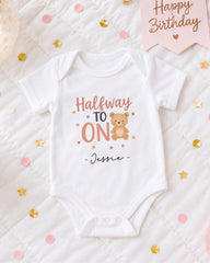 A white onesie with a teddy bear and the text "Halfway to ON" printed on it, along with the name " Jessie" written below. The onesie is placed on a bed with confetti and a birthday card.