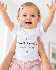 A smiling baby girl wearing a white t-shirt with text and a heart, being held up by someone's arms.