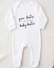 A white onesie with the text "your bestie is growing a baby bestie" printed on it.