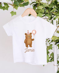 A white t-shirt with a cartoon image of a bear wearing a party hat and holding a balloon, along with the name "Jamey" printed on it.
