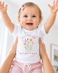 A smiling baby girl wearing a white t-shirt with a floral design and the text "One Little Wildflower" is being held up by someone's arms.