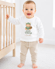 A smiling baby boy wearing a white sweatshirt with a Viking graphic and the text "Little Viking" is standing next to a wooden crib.