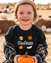 A young child wearing a black t-shirt with a pumpkin and bat design, smiling and holding a small orange pumpkin.