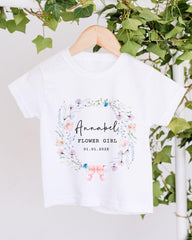 A white t-shirt with a floral wreath design and text printed on it, hanging on a wooden hanger.
