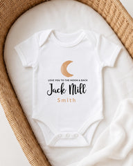 A white onesie with a crescent moon and the text "Love you to the moon & back Jack Mill Smith" printed on it, laid out on a white blanket in a basket.
