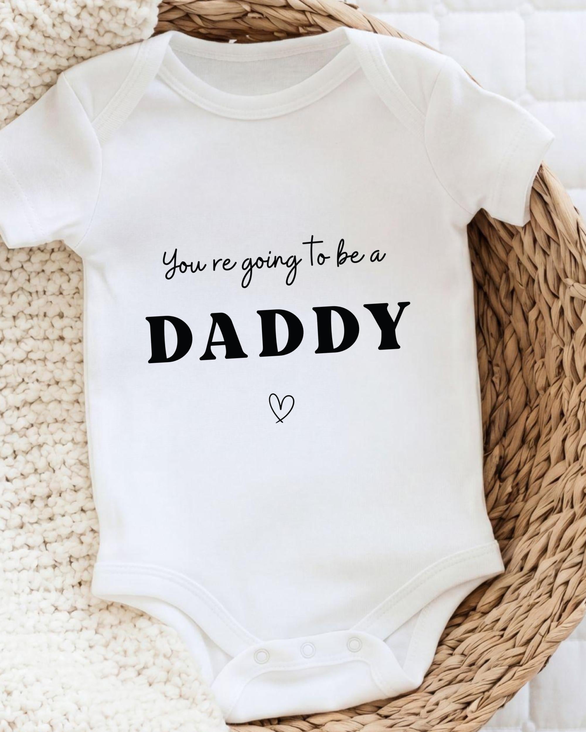 A white baby onesie with the text "You're going to be a DADDY" printed on it, along with a small heart symbol.