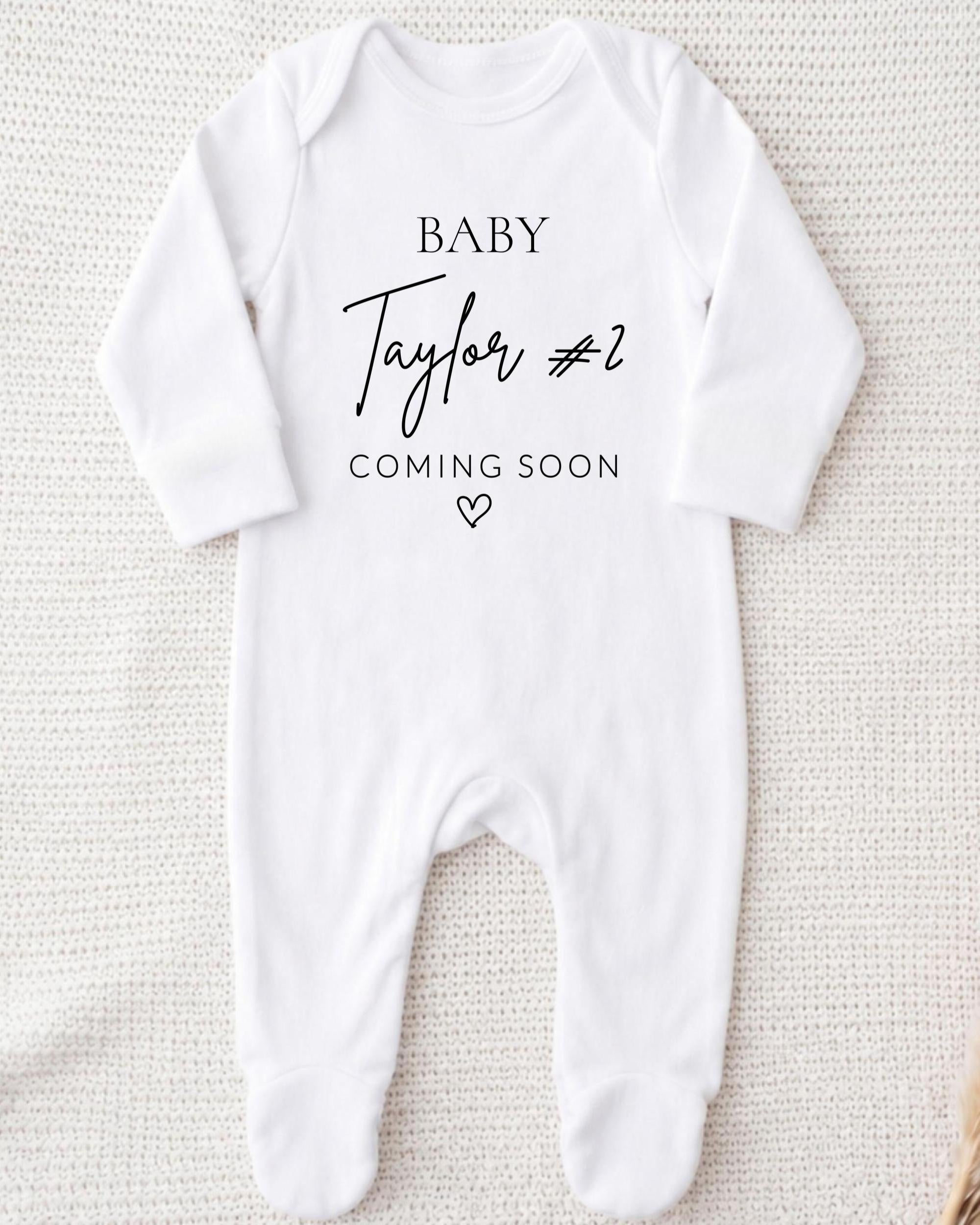 A white onesie with the text "BABY Taylor #2 COMING SOON" printed on it.