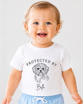 A smiling baby wearing a white t-shirt with a graphic of a dog and the text "PROTECTED BY Bella" printed on it.