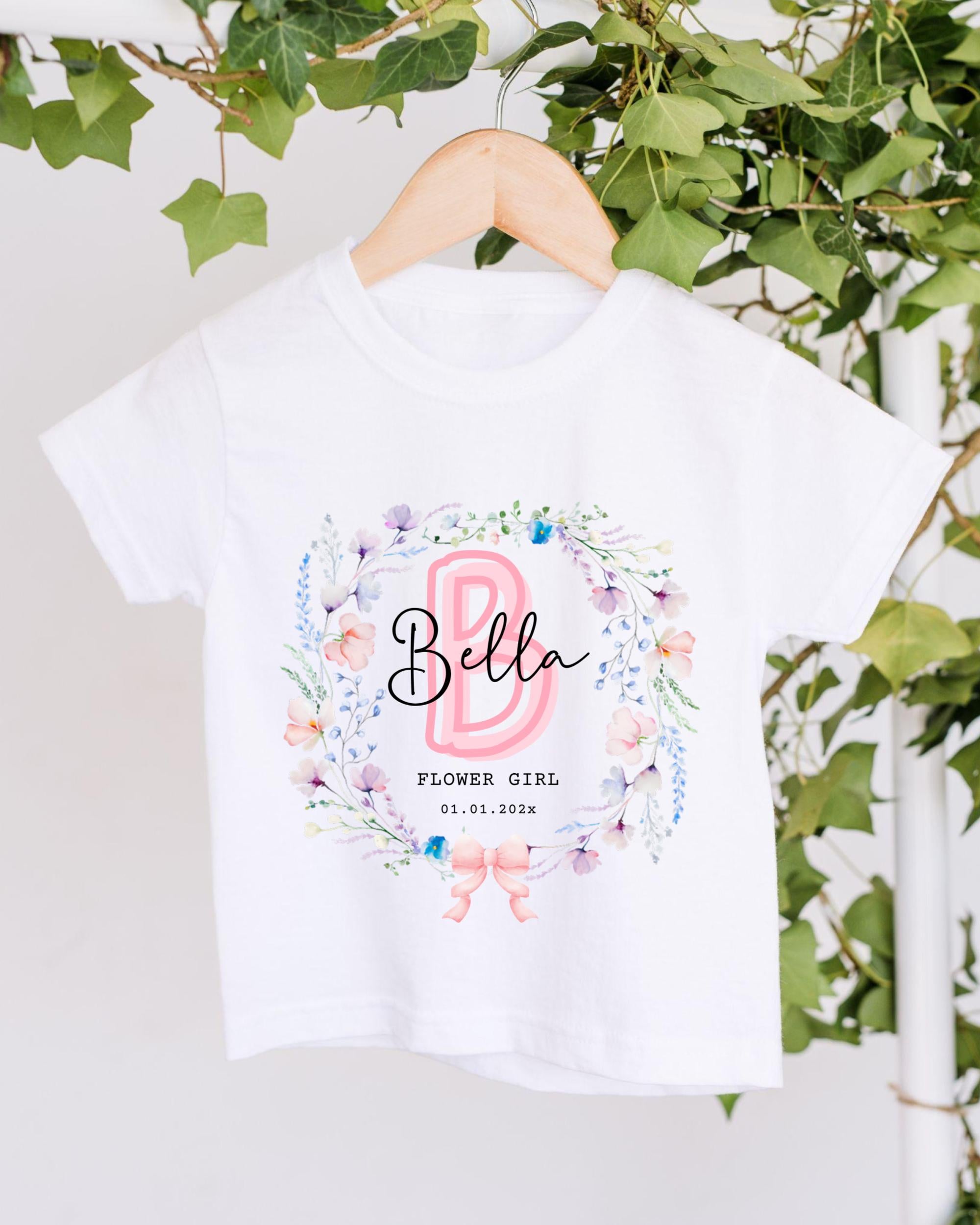 A white t-shirt with a floral wreath design and the text "Bella" printed on it, hanging on a wooden hanger.
