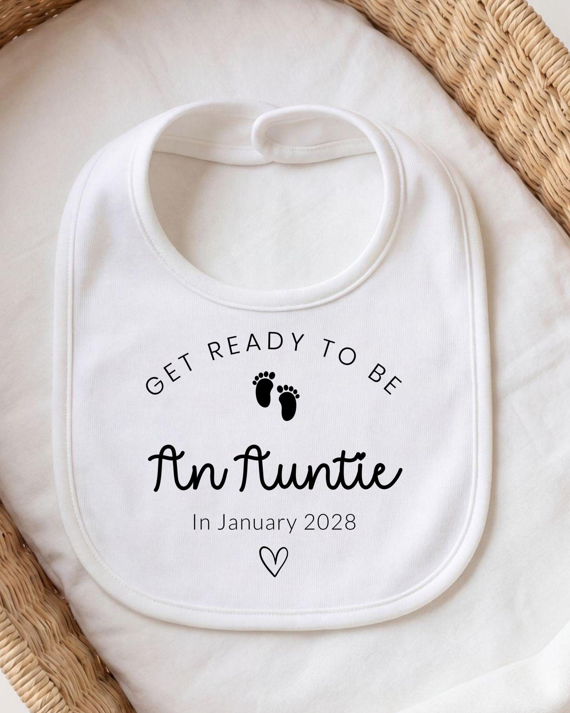 A white bib with the text "GET READY TO BE An Auntie In January 2028" printed on it, along with a small black footprint symbol.