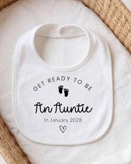 A white bib with the text "GET READY TO BE An Auntie In January 2028" printed on it, along with a small black footprint symbol.