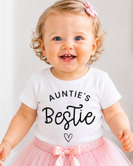 A smiling baby girl wearing a white t-shirt with the text "Auntie's Bestie" and a pink tutu skirt.