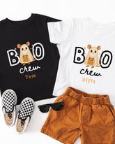 A pair of children's clothing, including a black t-shirt with a ghost character and the text "BOO crew Tom Alfie", a white t-shirt with the same text, and a pair of brown shorts. There are also a pair of black and white checkered shoes and a pair of sunglasses on the floor.