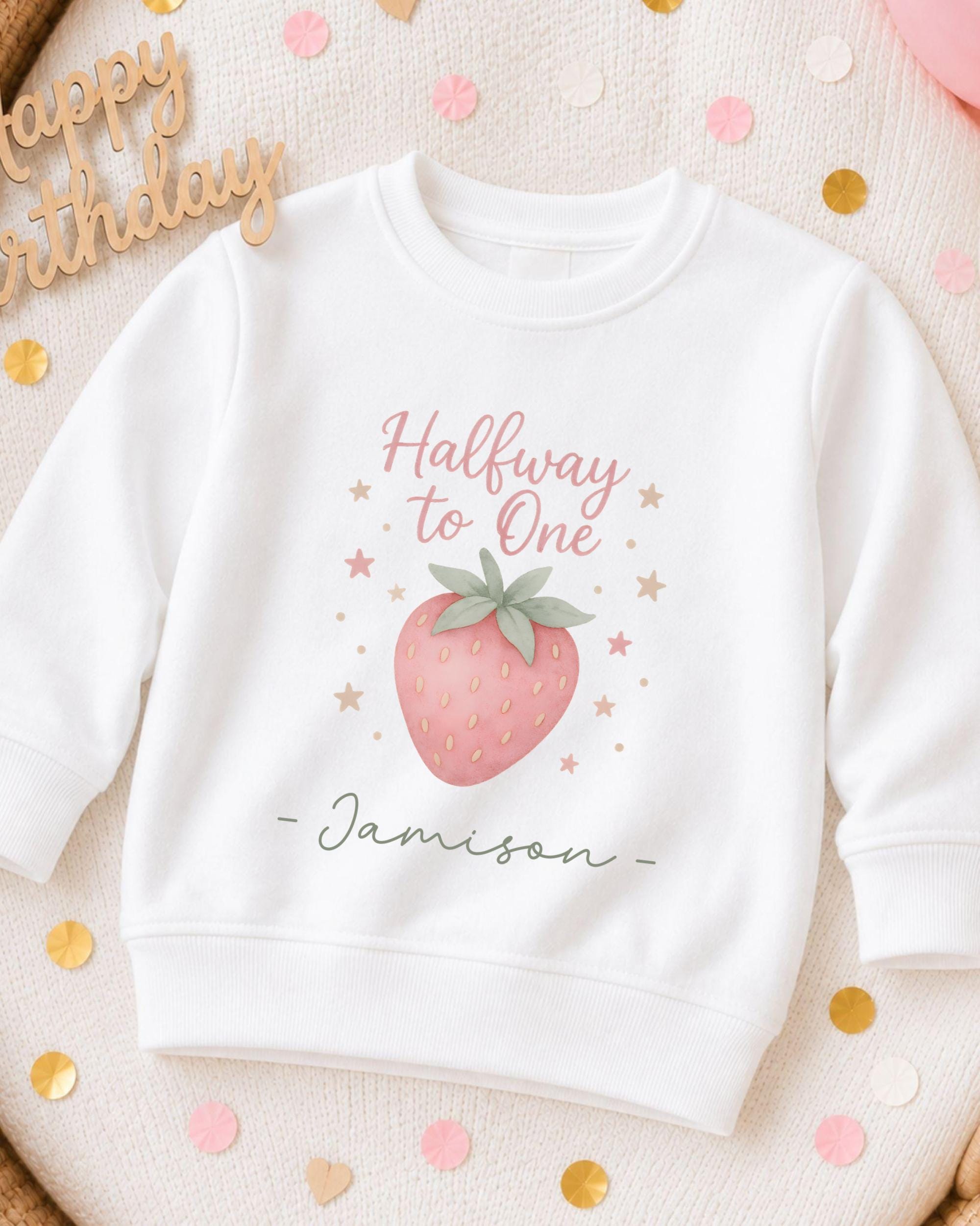 A white sweatshirt with a pink strawberry graphic and the text "Halfway to One" printed on it, along with the name "Jasison" written below. The sweatshirt is surrounded by confetti.