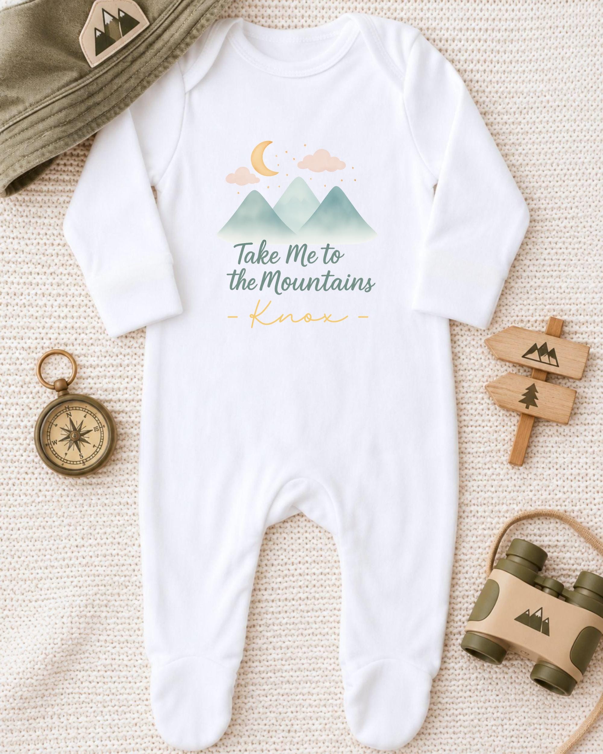 A baby onesie with a mountain and moon design, a compass, a hat, and a pair of binoculars are arranged on a beige surface.