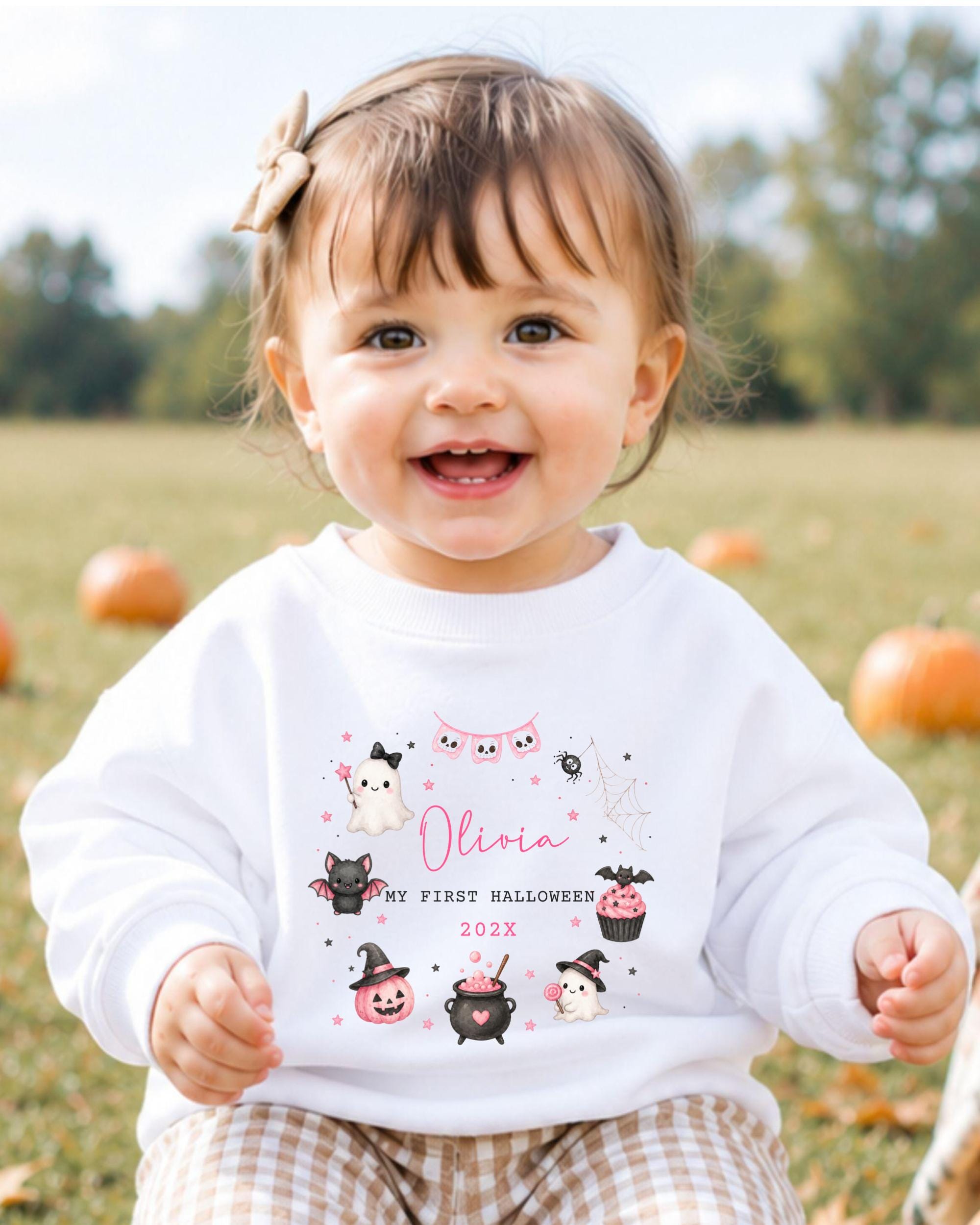 A smiling baby girl wearing a white sweater with a personalized design and text.