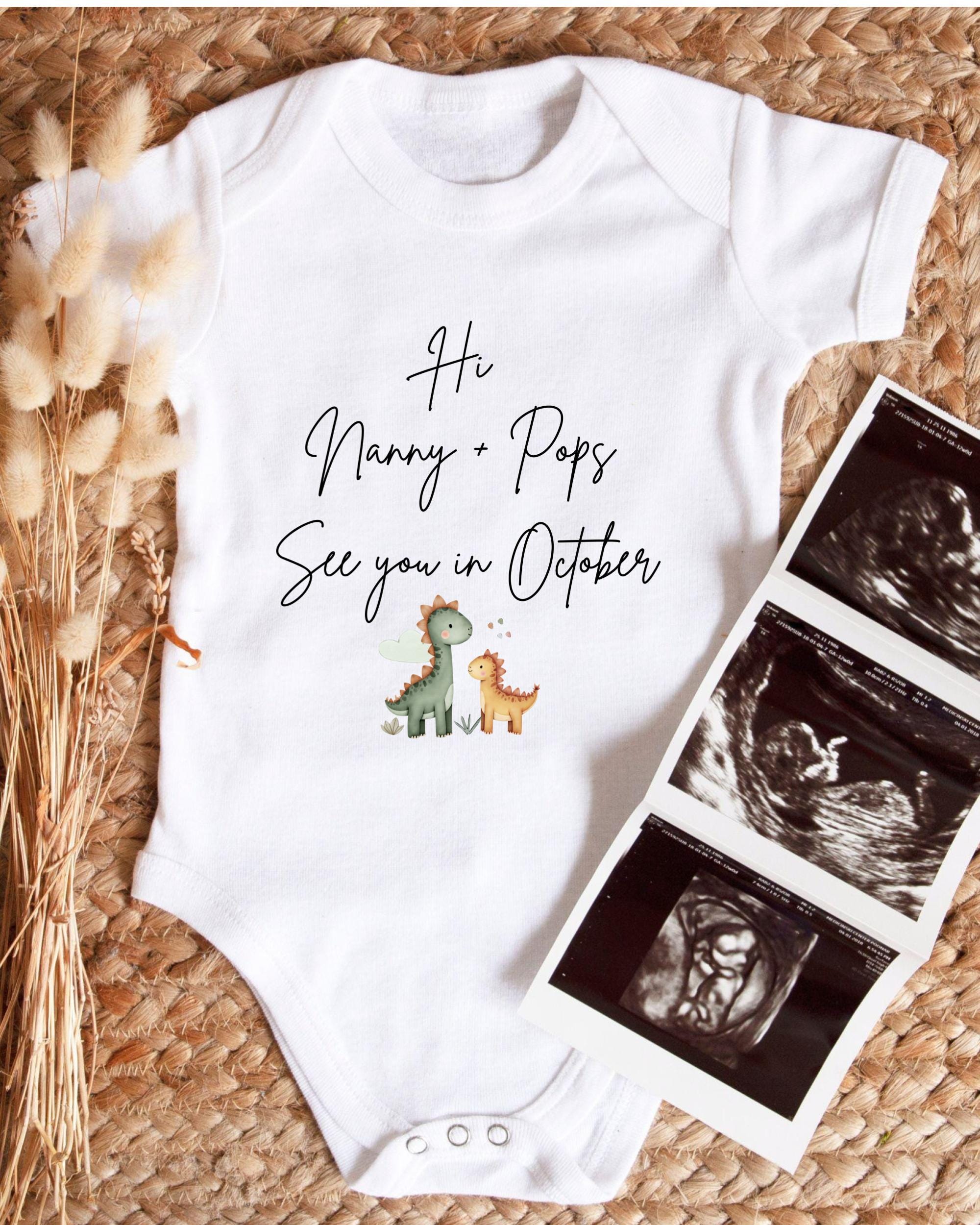 A white baby onesie with a dinosaur design and text that says "Hi Nanny + Pops See you in October" is displayed alongside a set of three black and white images, likely baby ultrasound images, on a woven surface.