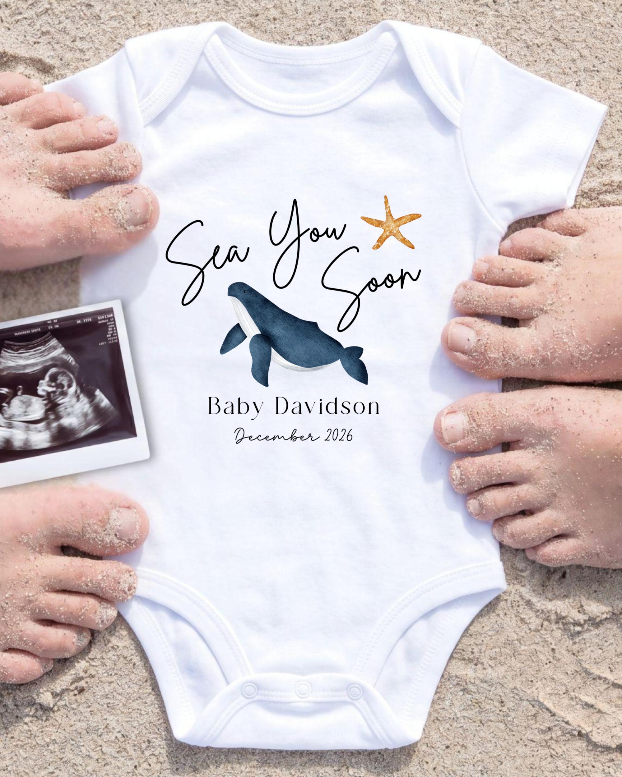 A white baby onesie with a blue whale and starfish design, surrounded by sand and a photograph of a baby's feet.