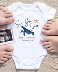 A white baby onesie with a blue whale and starfish design, surrounded by sand and a photograph of a baby's feet.
