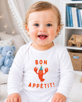 A smiling baby wearing a white onesie with a lobster and the text " bon appétit!" in front of a bookshelf.