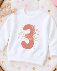 A white sweatshirt with the number 3 and the name "Hattie" printed on it, surrounded by pink and gold confetti.