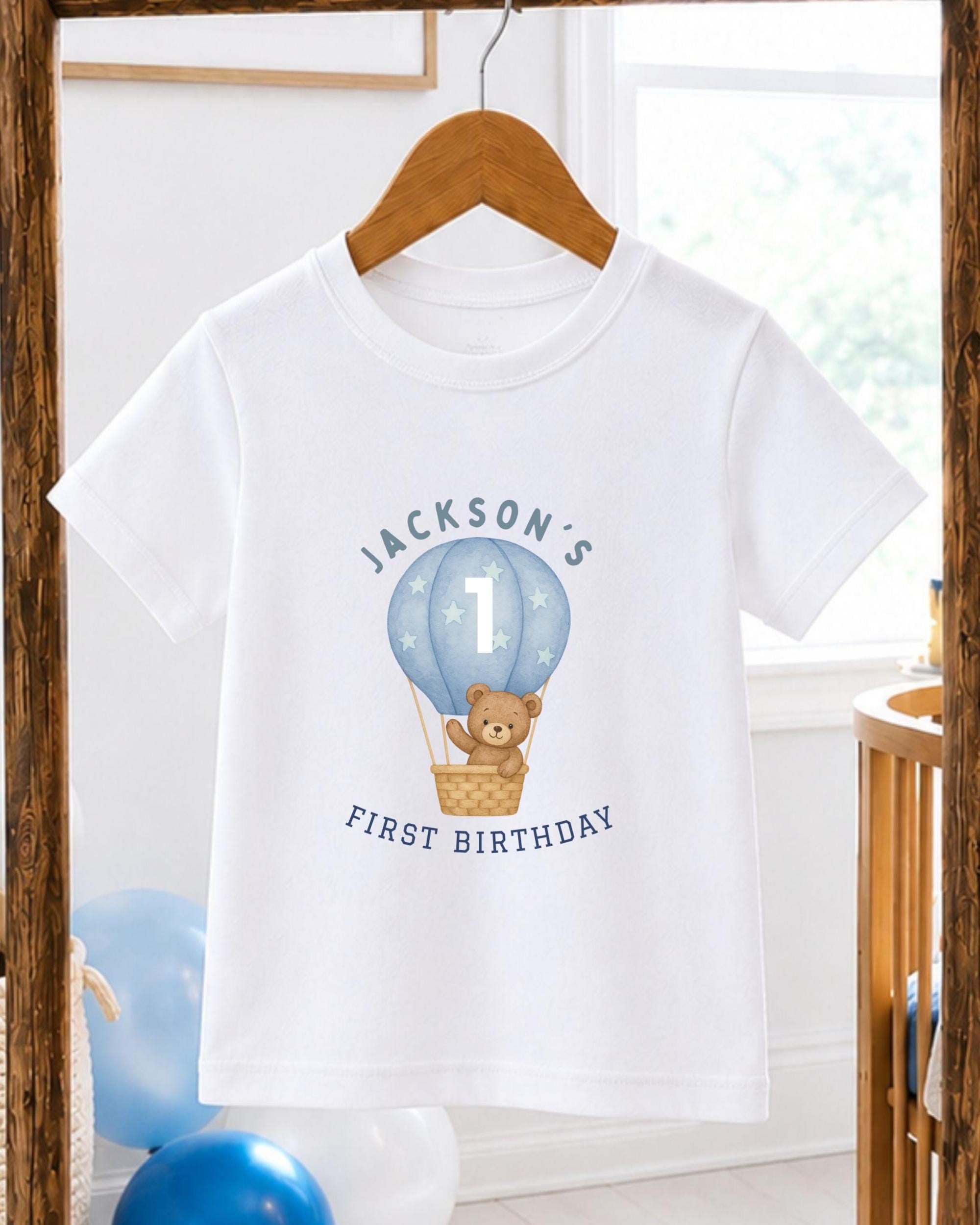 A white t-shirt with a graphic design of a teddy bear in a hot air balloon, along with the text "JACKSON'S FIRST BIRTHDAY".