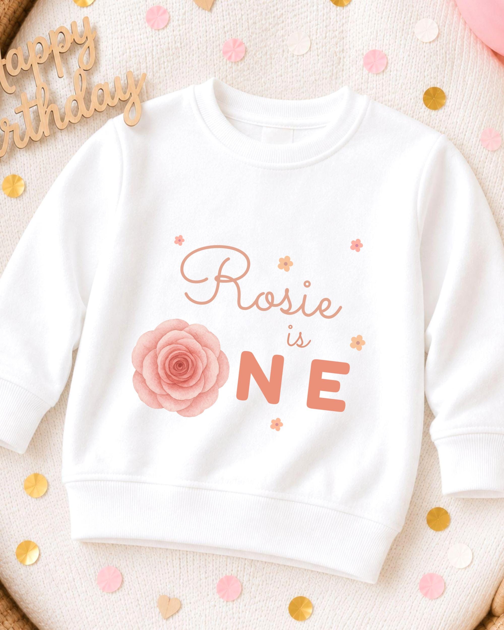 A white sweatshirt with a pink rose and the text "Rosa is ONE" printed on it, surrounded by confetti.