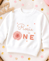 A white sweatshirt with a pink rose and the text "Rosa is ONE" printed on it, surrounded by confetti.