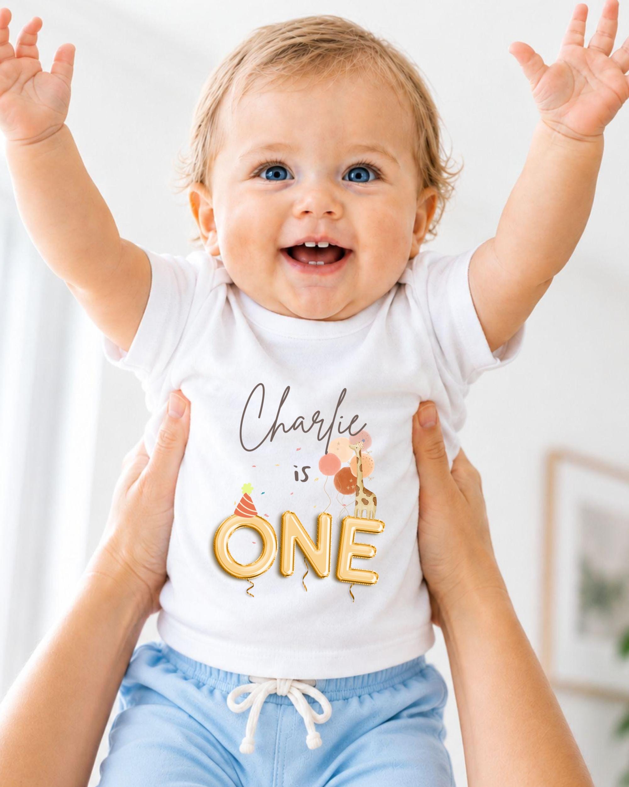 A smiling baby wearing a white t-shirt with the text "Charlie is ONE" is being held up by someone's arms.