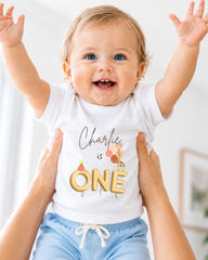 A smiling baby wearing a white t-shirt with the text "Charlie is ONE" is being held up by someone's arms.