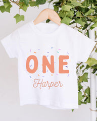 A white t-shirt with the text "ONE Harper" printed on it, along with colorful sprinkles. The t-shirt is hanging on a wooden hanger, and there are green leaves visible in the background.