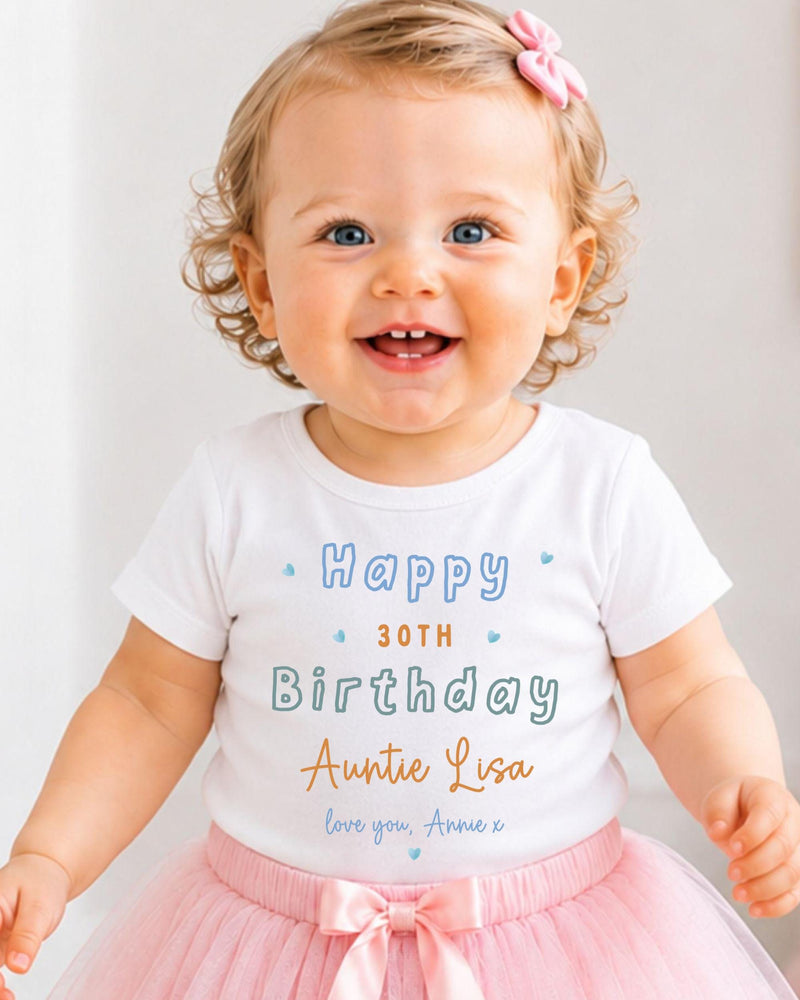 A smiling baby girl wearing a white t-shirt with text and a pink bow, standing in front of a white background.