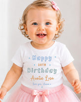 A smiling baby girl wearing a white t-shirt with text and a pink bow, standing in front of a white background.