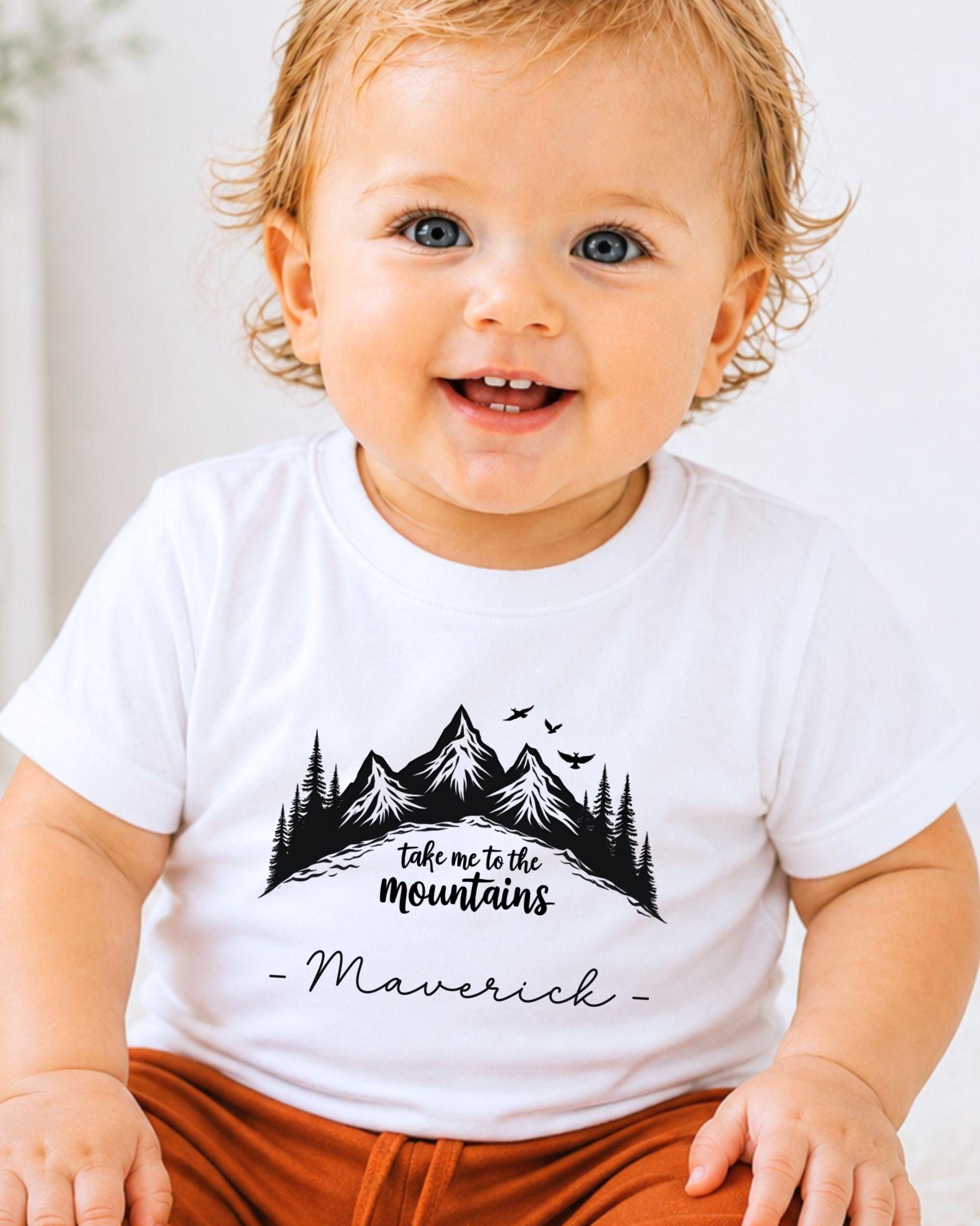 A smiling baby wearing a white t-shirt with a mountain graphic and the text "Take me to the mountains" and "Maverick" printed on it.