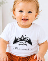 A smiling baby wearing a white t-shirt with a mountain graphic and the text "Take me to the mountains" and "Maverick" printed on it.