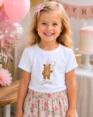 A young girl wearing a white t-shirt with a teddy bear and the number 4, smiling at the camera, with pink balloons and a birthday cake in the background.