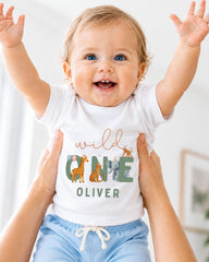 A smiling baby wearing a white t-shirt with a graphic of animals and the name "Olivier" printed on it, being held up by someone's arms.