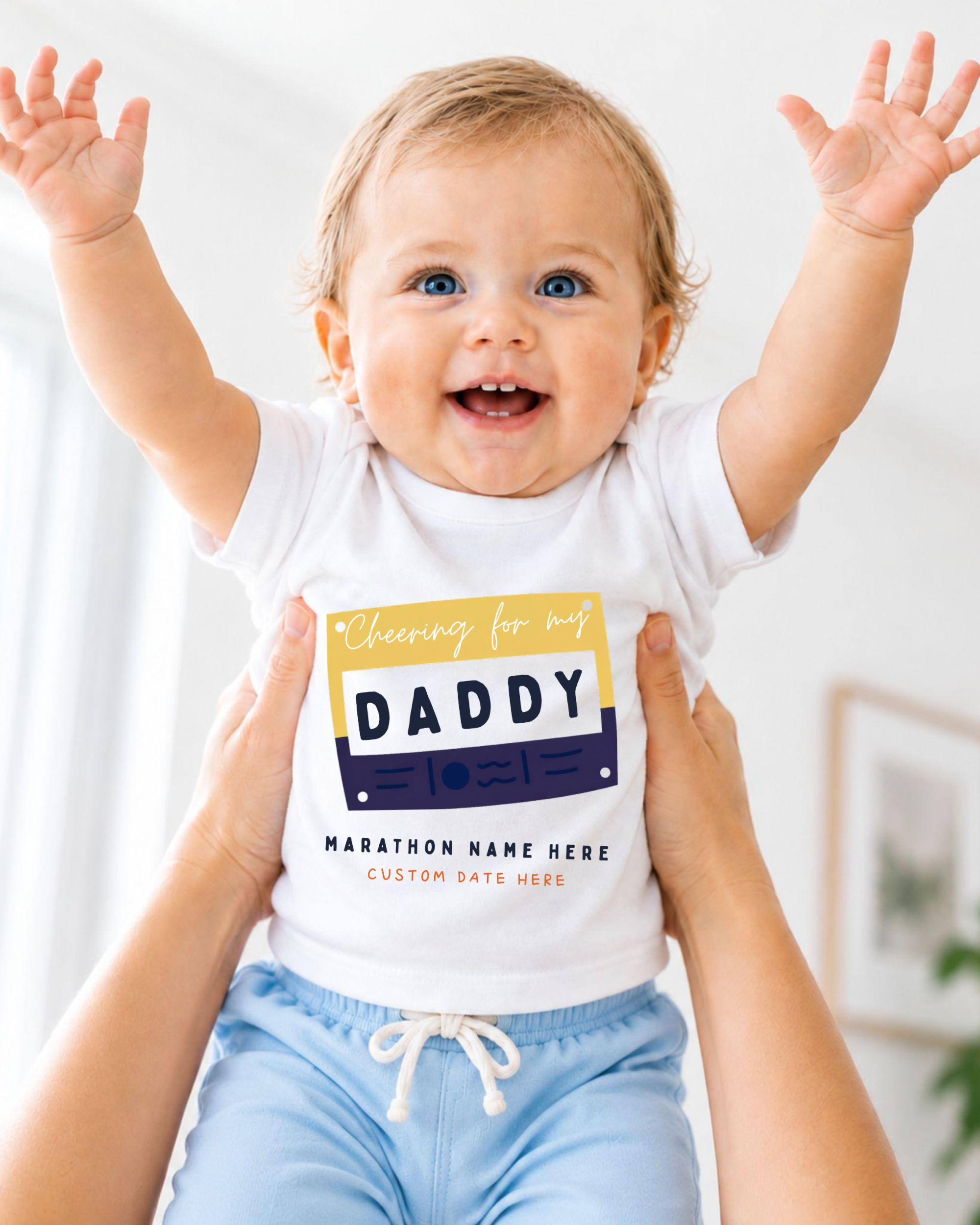 A smiling baby wearing a white shirt with a graphic and text, being held up by someone's arms.