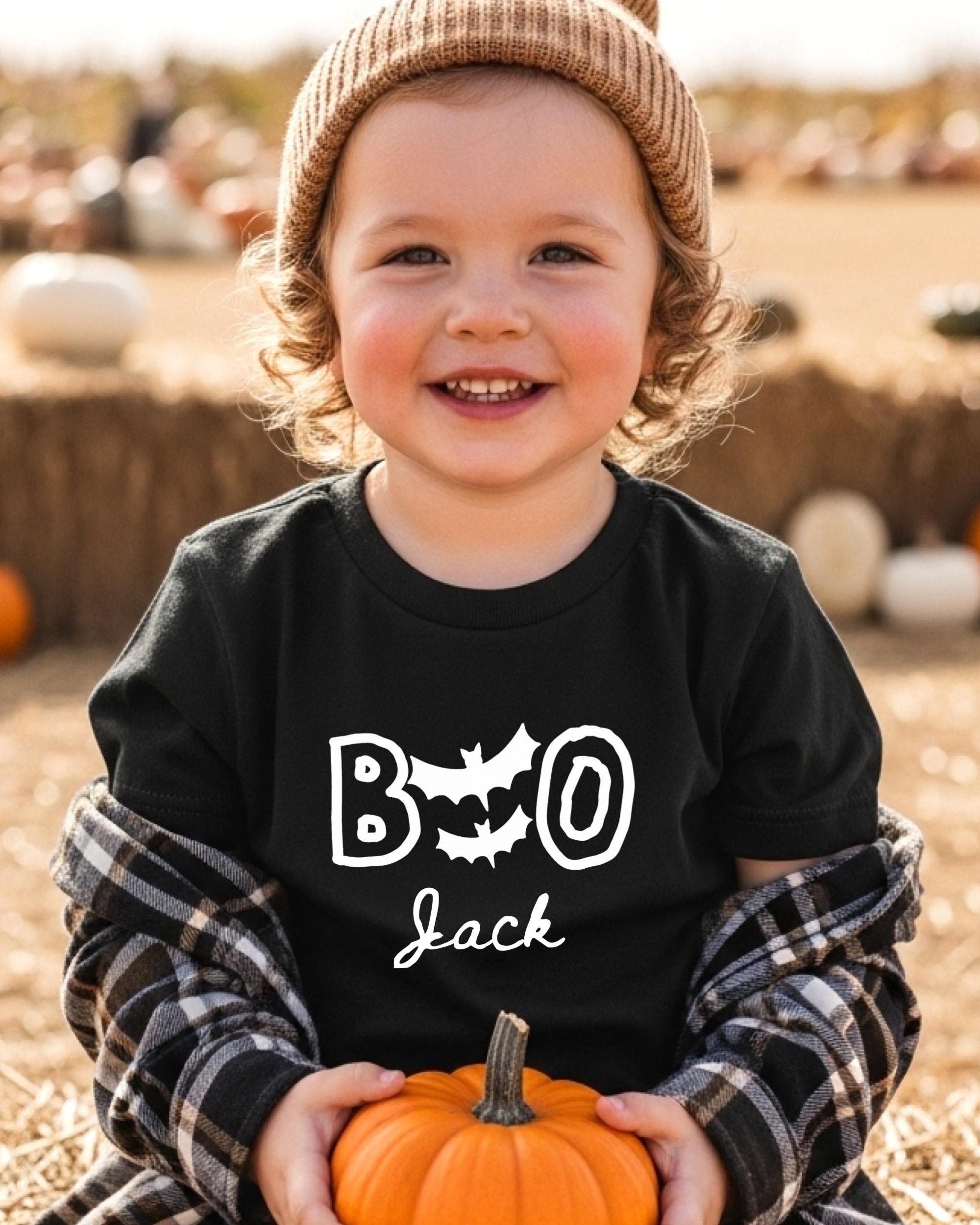 A young child wearing a black t-shirt with the text "BOO Jack" and a beanie is holding a small orange pumpkin.