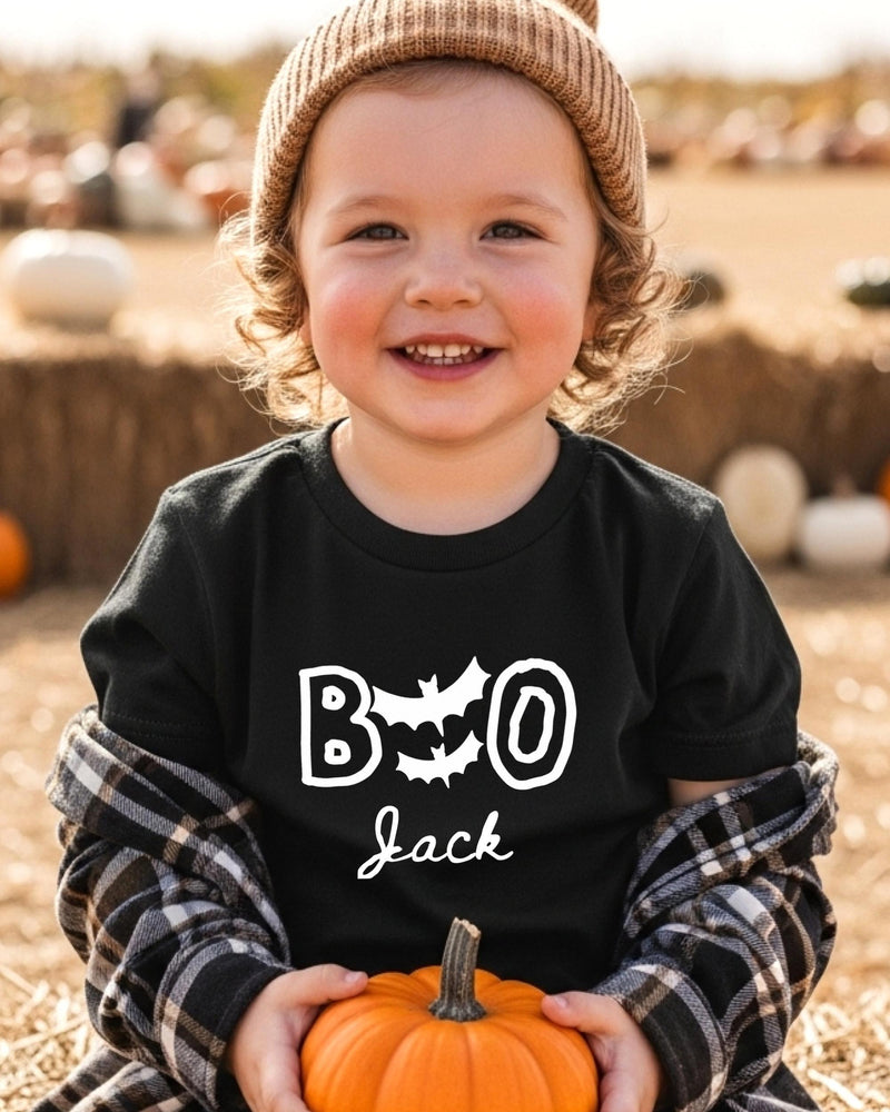 A young child wearing a black t-shirt with the text "BOO Jack" and a beanie is holding a small orange pumpkin.