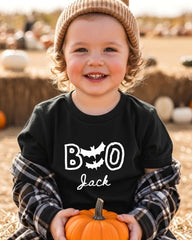 A young child wearing a black t-shirt with the text "BOO Jack" and a beanie is holding a small orange pumpkin.